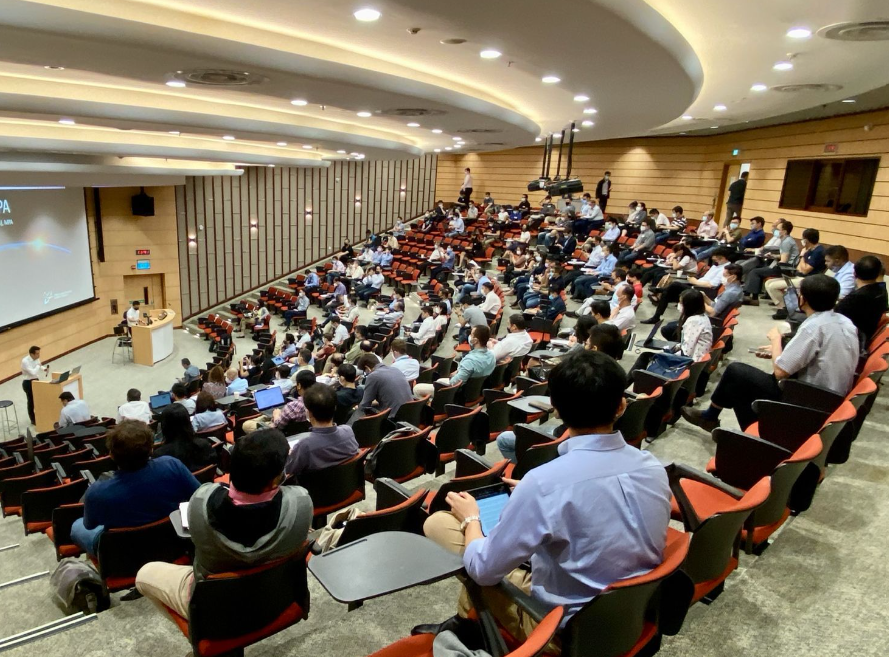 A lecture hall filled with people sitting in tiered seats, listening to a speaker at a podium near a large screen.